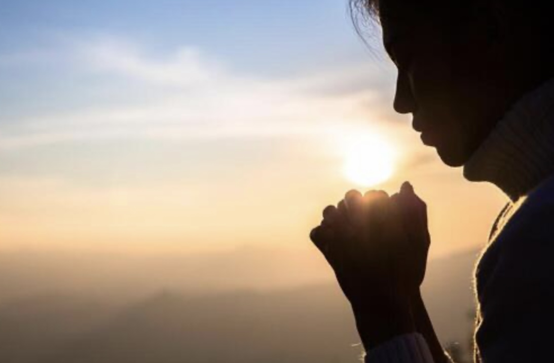 A woman in silhouette, hands joined in prayer, facing the rising sun