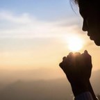 A woman in silhouette, hands joined in prayer, facing the rising sun